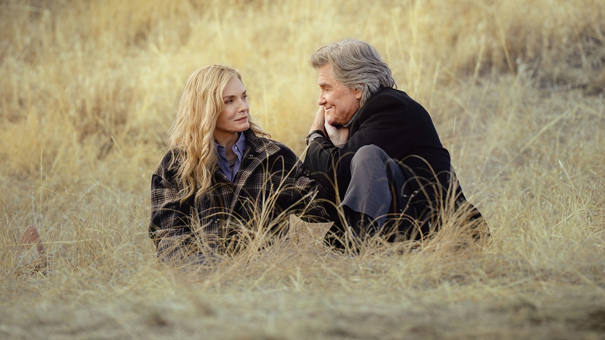 Michelle Pfeiffer and Kurt Russell sit together in the long dry grass facing each other during a quiet time outside.