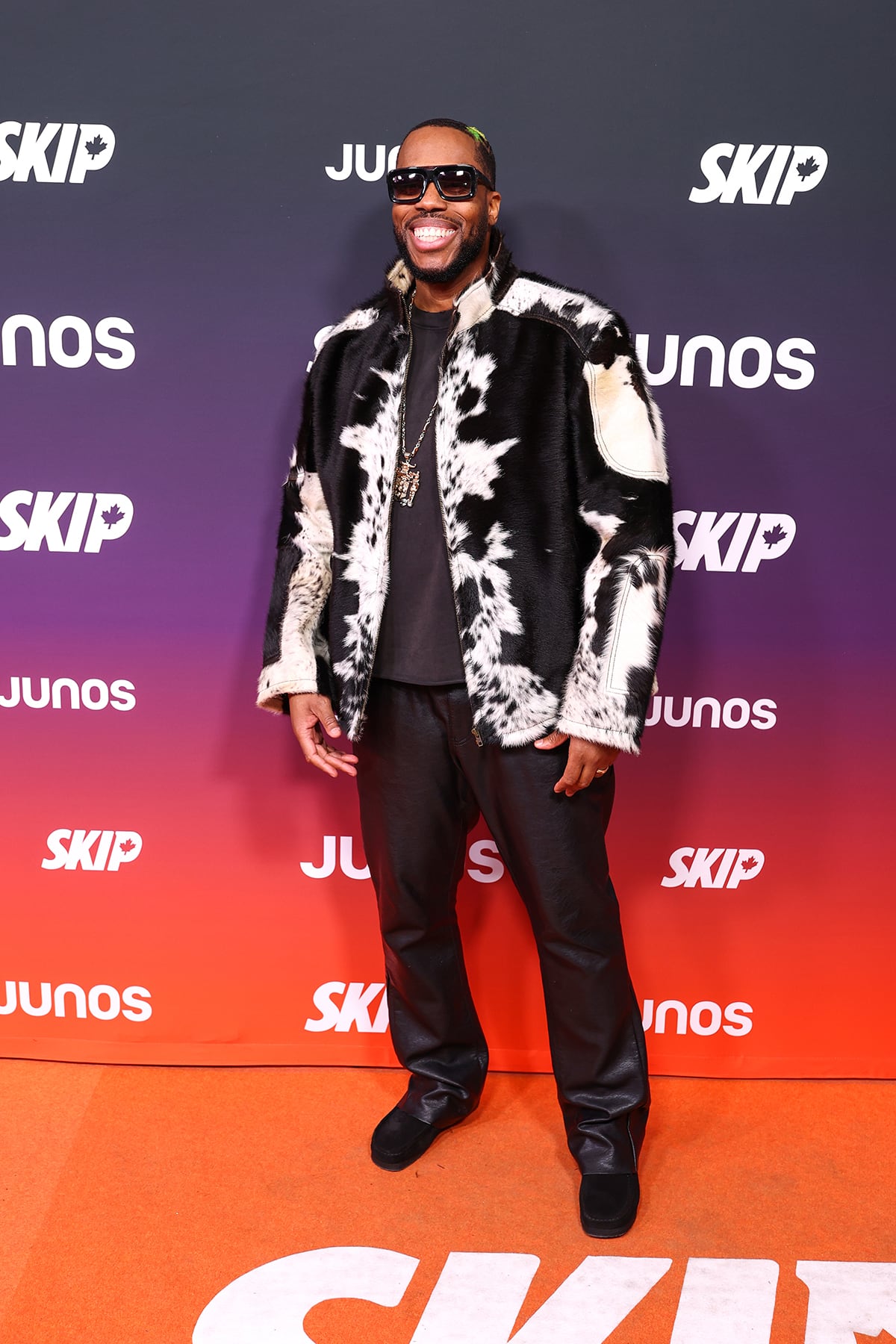 A man is standing in a cow print jacket on an orange carpet with leather pants, oversized sunglasses and a large dangling necklace.