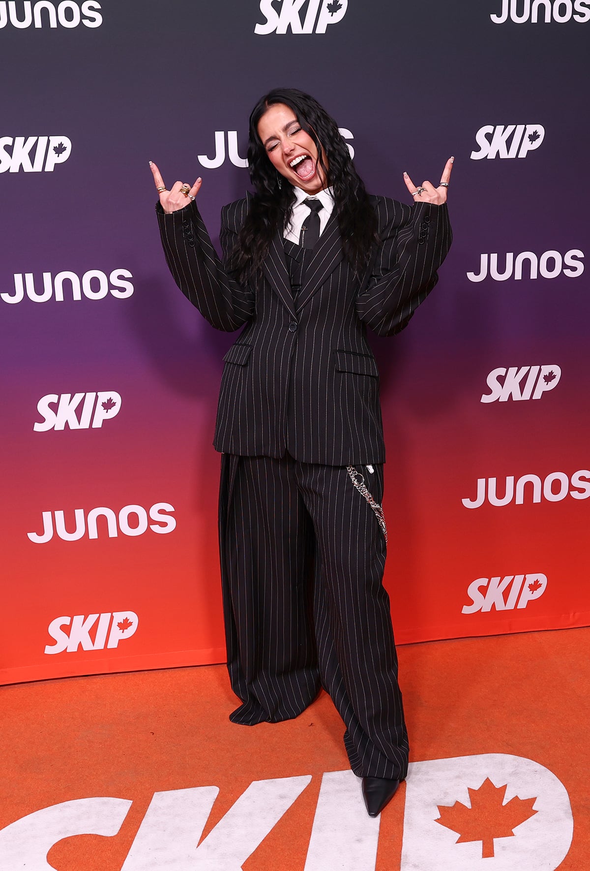 A woman stands with devil's horn arms raised and wearing an oversized pinstripe suit.