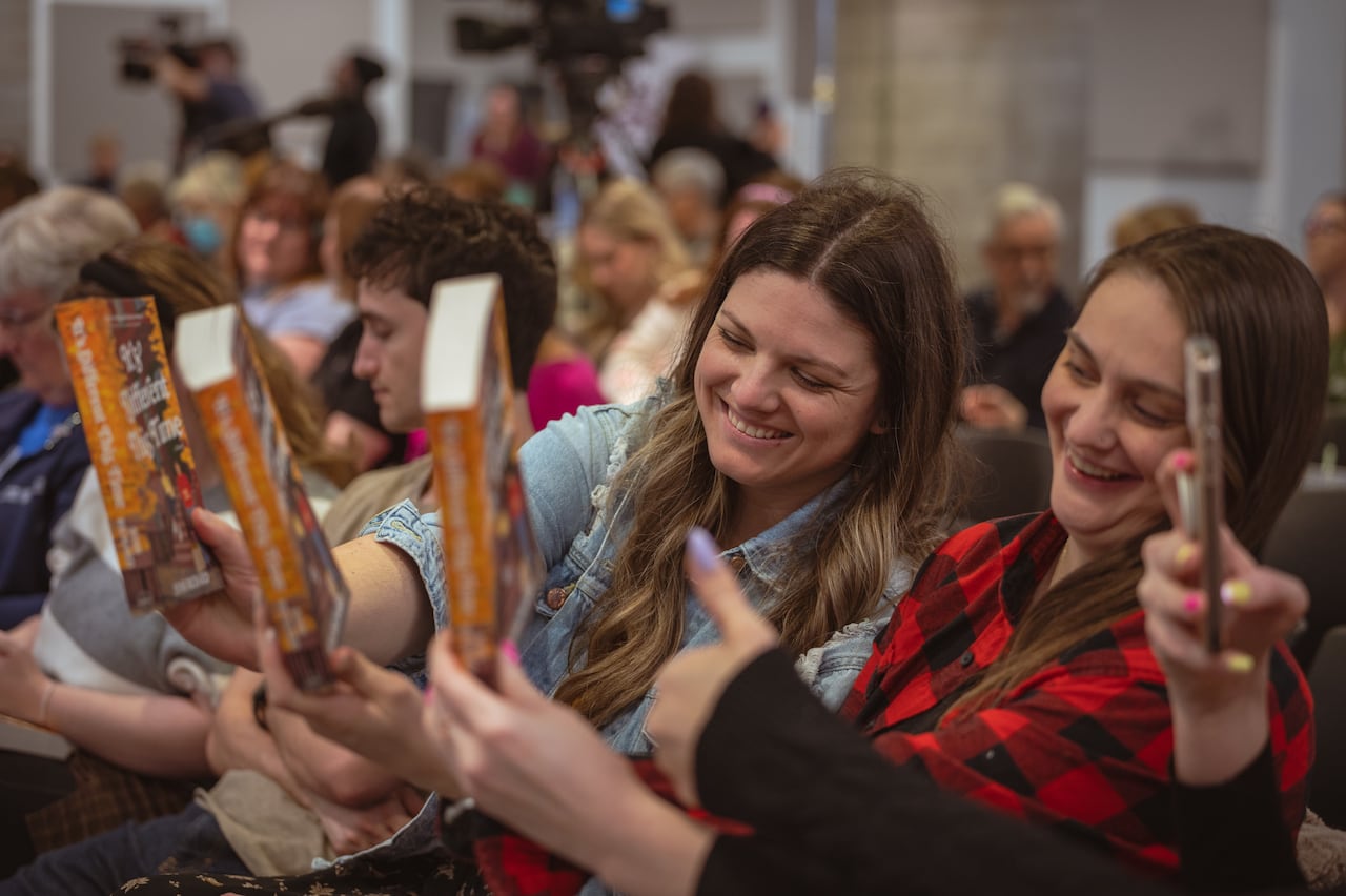 Three women hold up books and one of them takes a photo of them with a smartphone.