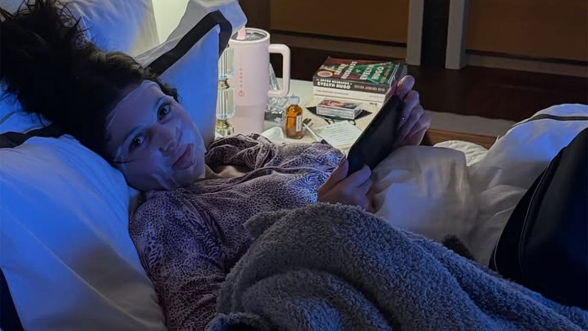 Kylie Jenner in a dark hotel room, with blue lights, wearing animal pajamas and a white face mask. He is holding an iPad and smiling widely at the camera. A table with drinks and books is near him.