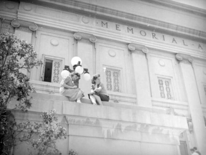 Black and white image of a Roman temple style building. Two women are sitting on a concrete pit. 