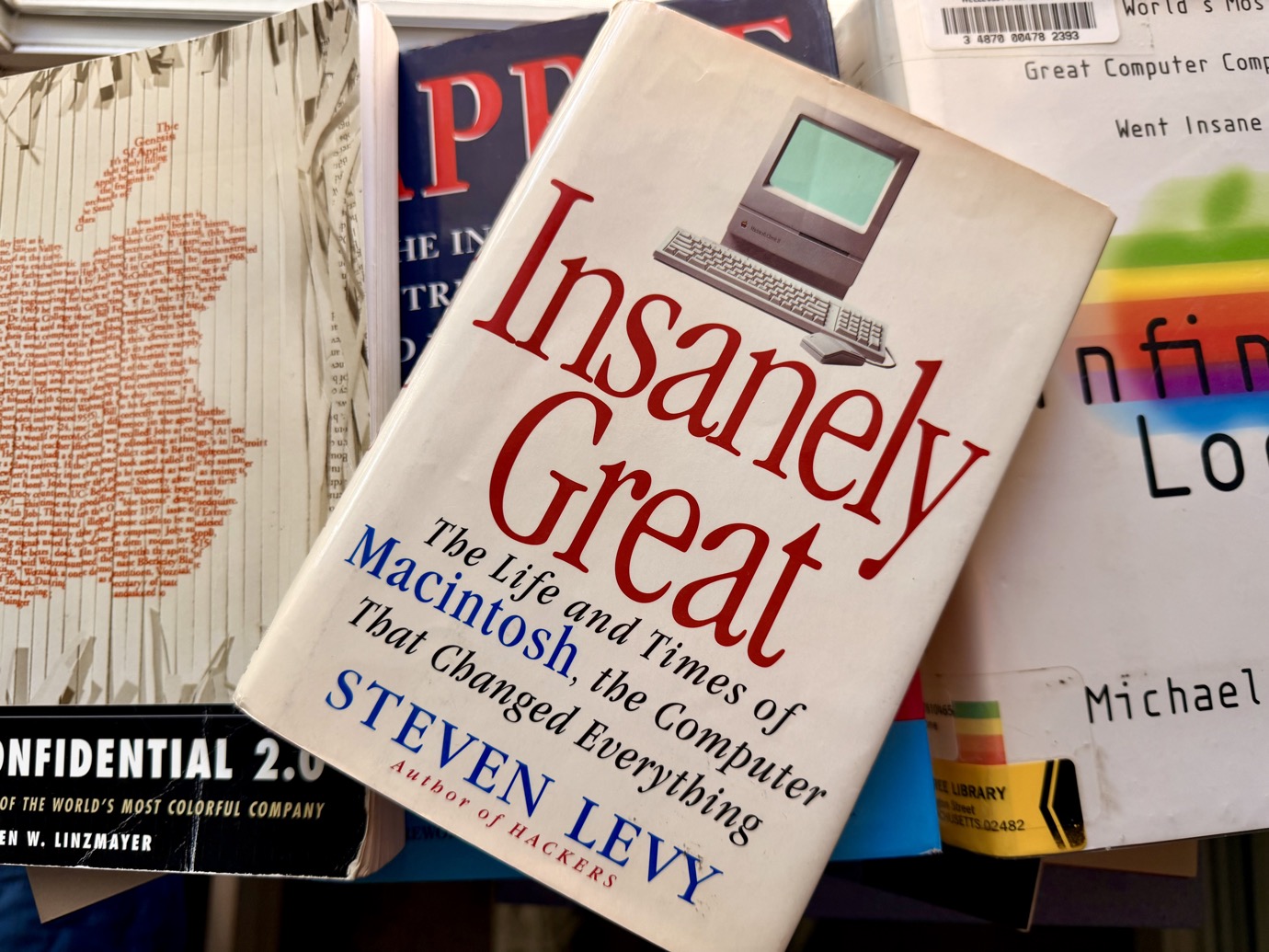 A book titled 'Insanely Great: The Life and Times of Macintosh, the Computer That Changed Everything' by Steven Levy, which has a classic computer analogy, is featured prominently among other books.