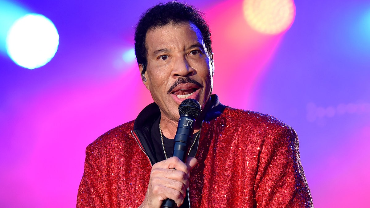 Lionel Richie speaks into a microphone on stage wearing a sparkly red jacket