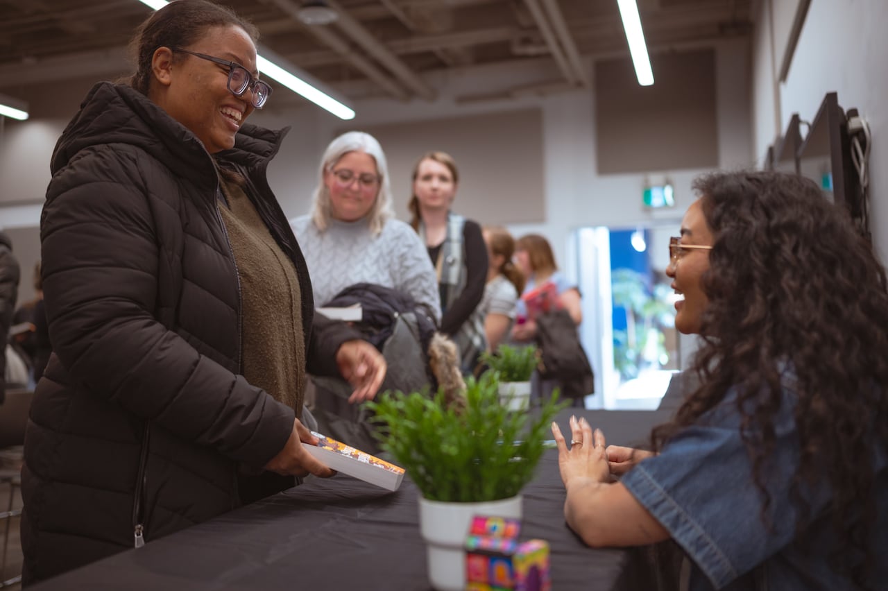 A smiling woman holding a book in front of another smiling woman sitting down at a table.